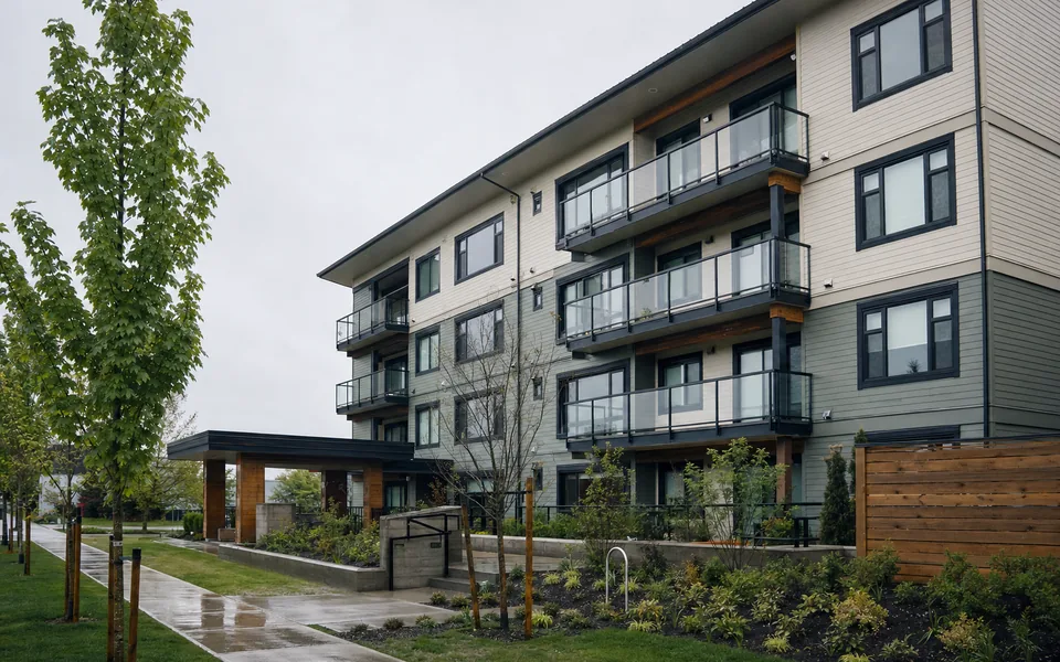 A four-storey wood-frame apartment building with balconies and grey and cream siding beside a wet path and young street trees (AI-generated image)