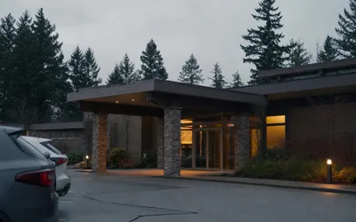 A low modern building with stone pillars and lit glass doors at dusk, parked cars and evergreen trees (AI-generated image)