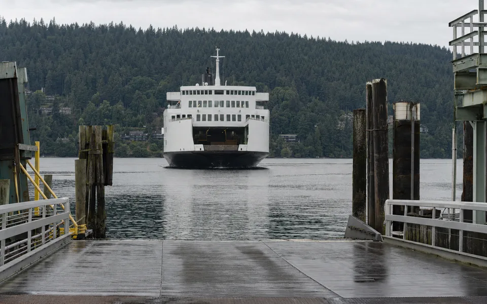 A wet steel vehicle ramp and timber pilings at a ferry berth, a white car ferry approaching across a calm bay below forested hills (AI-generated image)