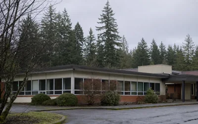 A low, flat-roofed 1970s school building with a long band of windows, an empty wet parking lot and tall firs (AI-generated image)