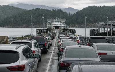 Rows of cars waiting on a wet ferry ramp toward an approaching white car ferry, forested hills behind (AI-generated image)