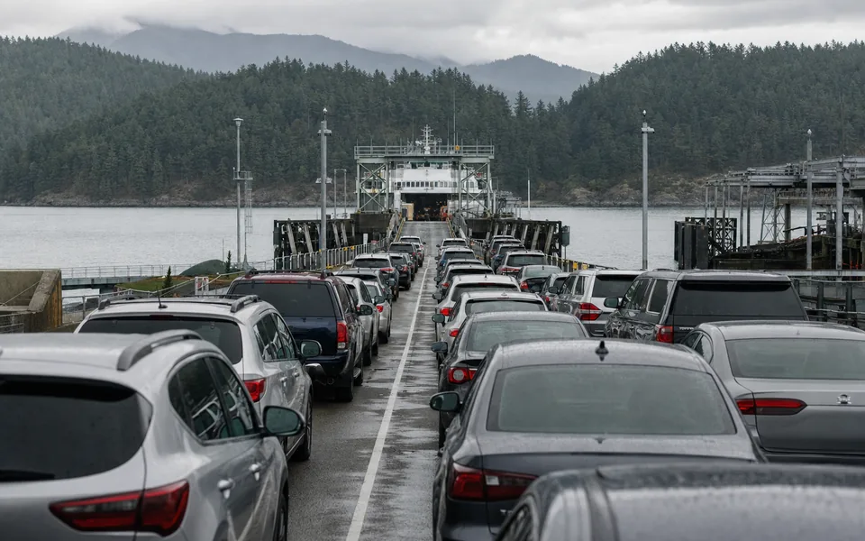 Rows of cars waiting on a wet ferry ramp toward an approaching white car ferry, forested hills behind (AI-generated image)