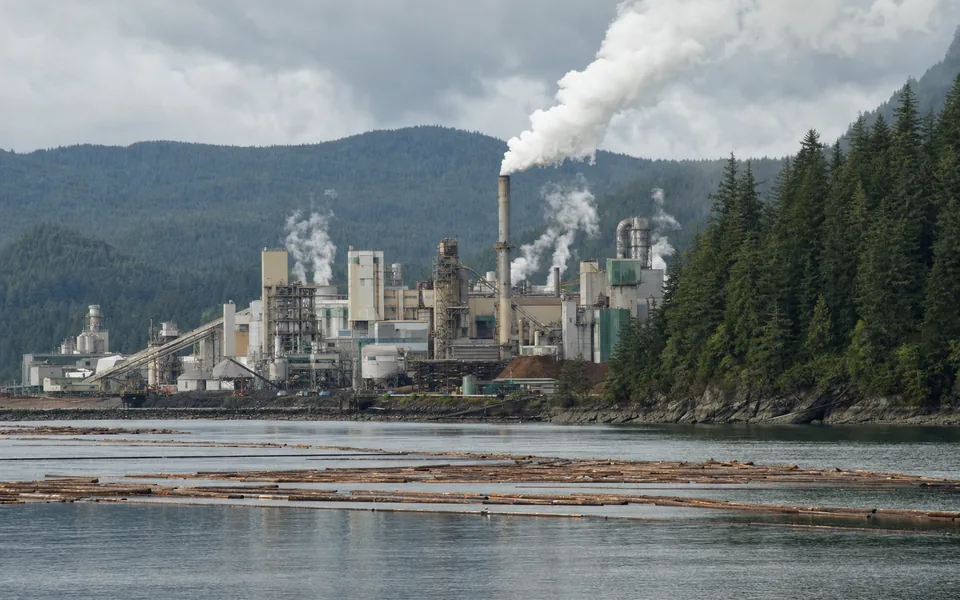 A large pulp mill with steam plumes on a forested shoreline, log booms floating in front (AI-generated image)