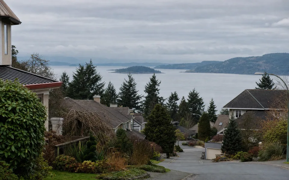 A hillside residential street of single-family West Coast houses looking down to the sea and islands under grey sky (AI-generated image)