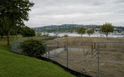 A chain-link-fenced excavated empty lot beside a grassy park slope, a harbour with moored boats beyond (AI-generated image)