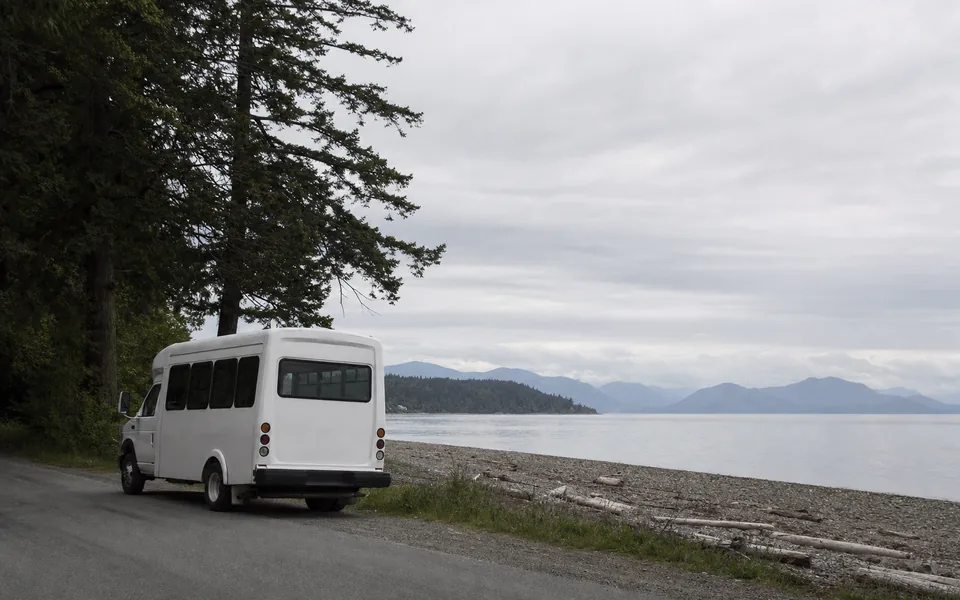 A plain white community minibus parked on a seaside road beside a pebble beach with the strait and mountains beyond (AI-generated image)