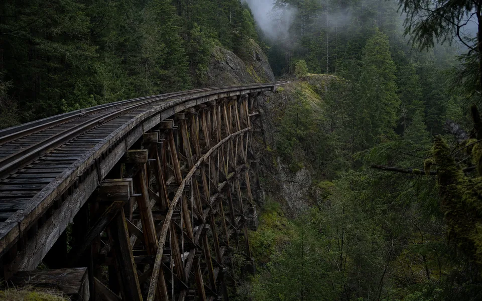 A single-track railway line curving over a tall timber trestle across a misty forested ravine (AI-generated image)