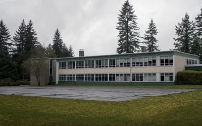 A two-storey school block with ribbon windows, a wet empty playground and tall firs under grey sky (AI-generated image)